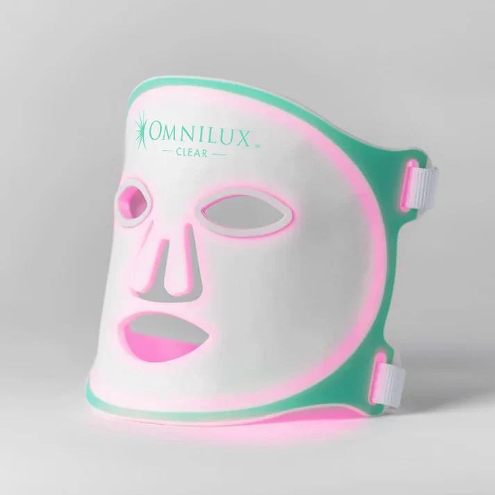 LED light therapy mask by Omnilux Clear designed for skincare. The mask features cutouts for eyes and mouth, with a sleek white and teal design. It emits pink light to promote clearer skin and reduce blemishes, suitable for home use. Ideal for achieving a radiant complexion, this device offers a non-invasive solution for various skin concerns. The adjustable straps ensure a comfortable fit for different face shapes. Perfect for those seeking to enhance their skincare routine with innovative technology.